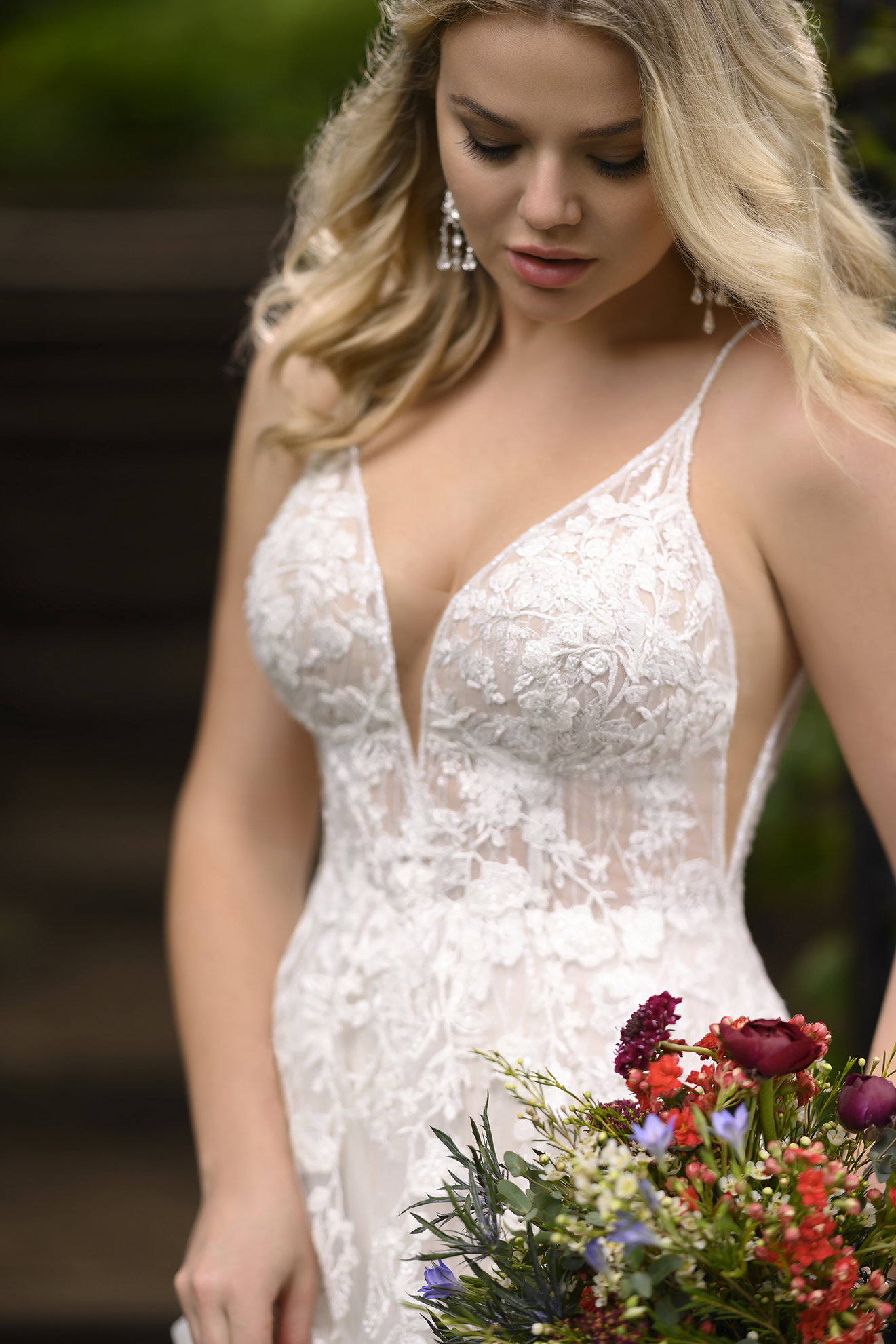 Romantisches Brautkleid aus Spitze mit tiefem Dekolleté und transparenten Einsätzen. Die Braut, die einen lebhaften Blumenstrauß trägt, blickt sanft nach unten und verstärkt so die zarte, elegante Anmutung des Hochzeitslooks.