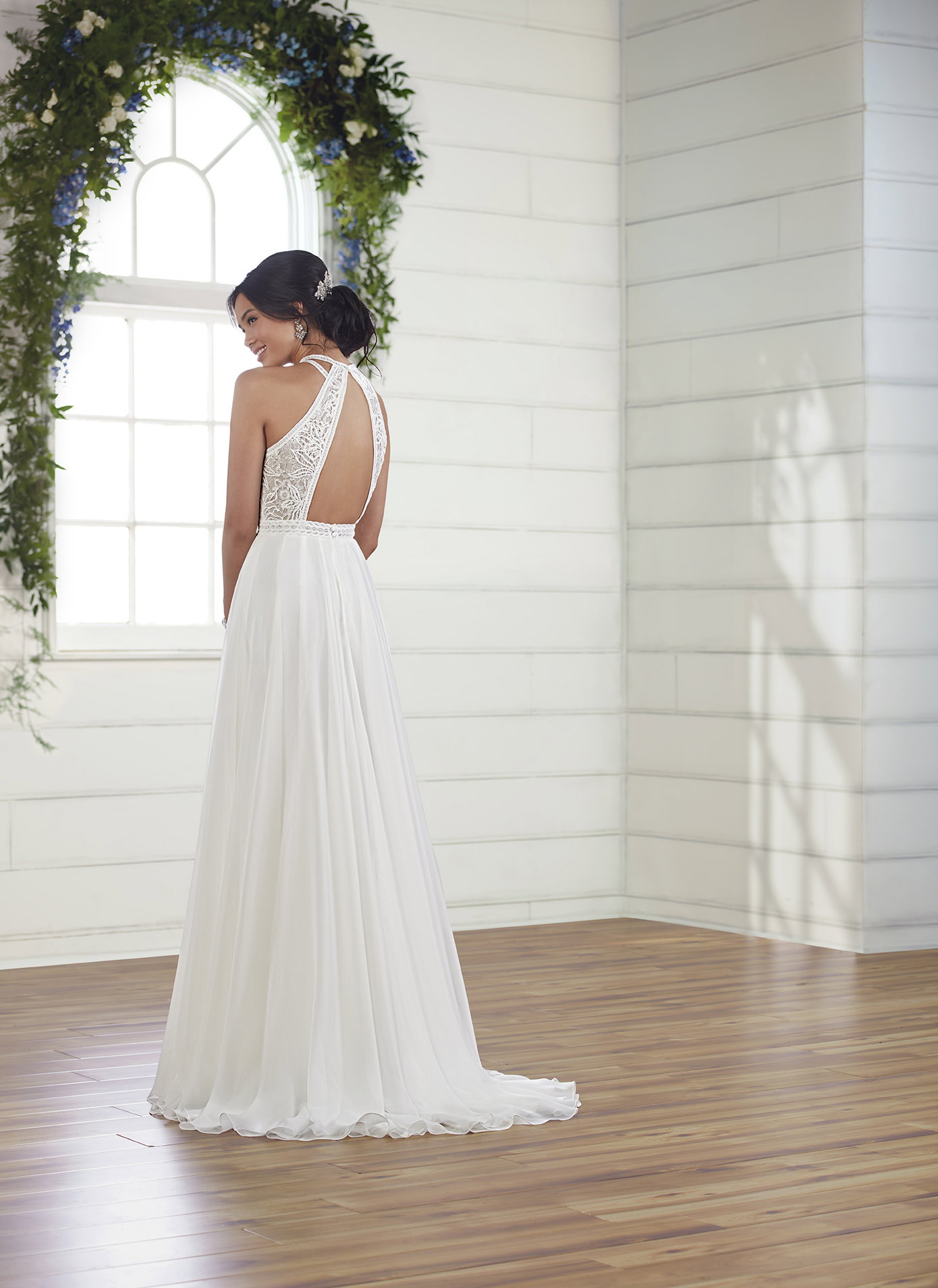 Eine Braut im schlichten, fließenden Kleid mit aufwendigem Spitzenrücken und elegantem Schnitt, steht in einem modernen, hellen Raum. Die grüne Dekoration über dem Fenster im Hintergrund setzt einen sanften, natürlichen Akzent.