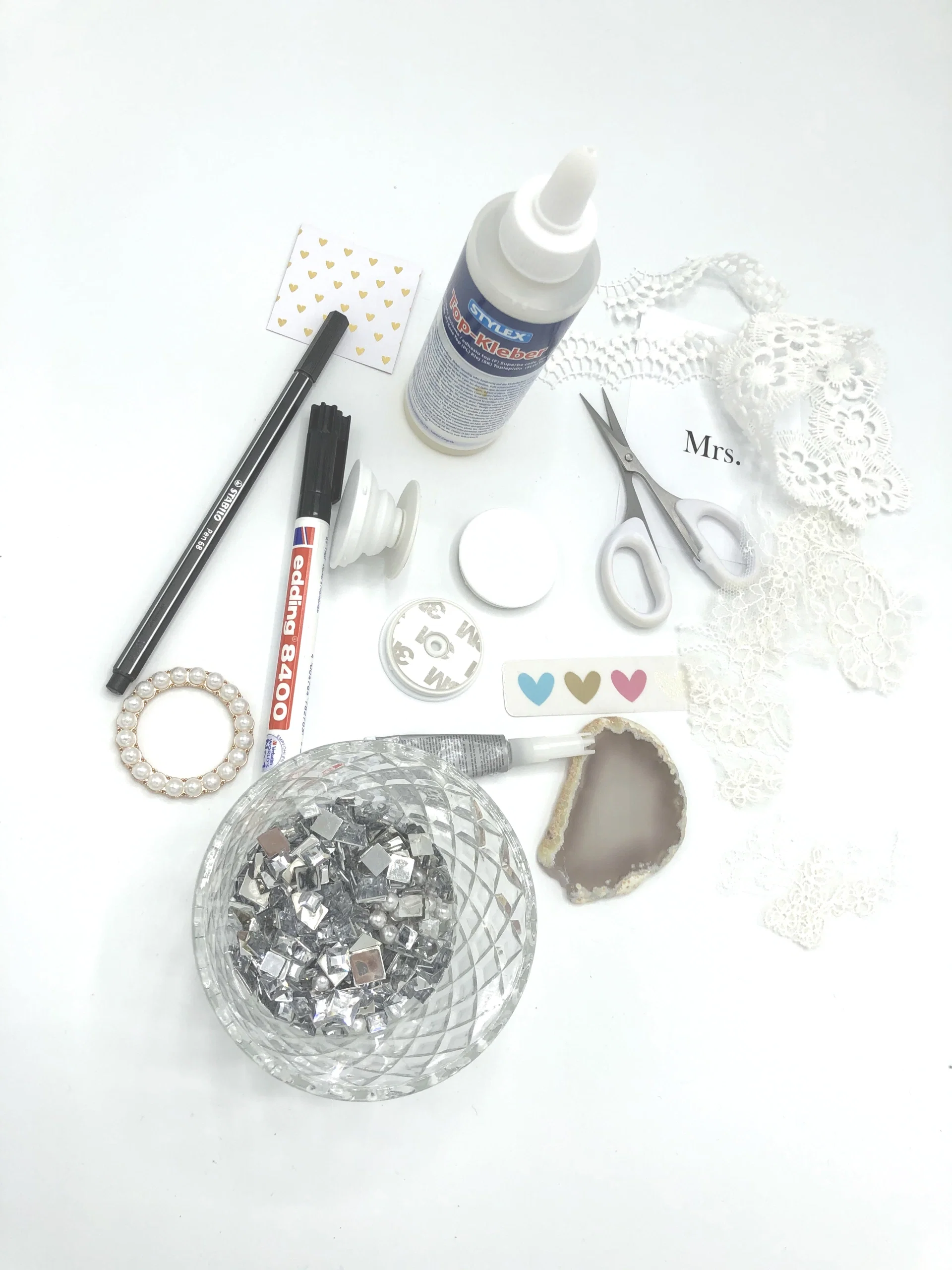 Verschiedene DIY-Materialien liegen bereit: Schere, Spitze, Kleber, Stifte und Glitzersteine. Ideal für die Gestaltung eines individuellen Popsockets.