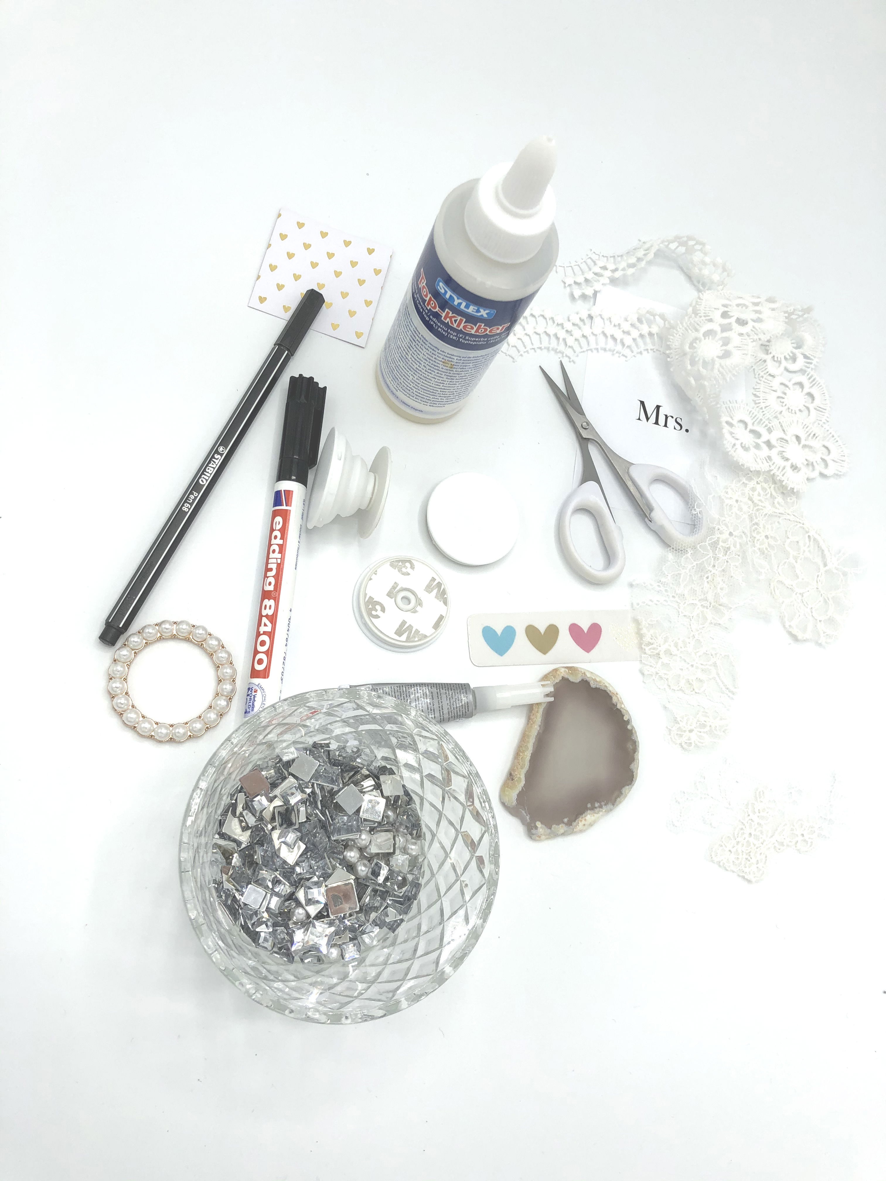 Verschiedene DIY-Materialien liegen bereit: Schere, Spitze, Kleber, Stifte und Glitzersteine. Ideal für die Gestaltung eines individuellen Popsockets.