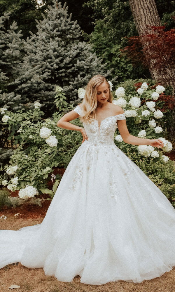 In einem eleganten Kleid mit Blumenmuster steht die Braut im Garten. Sie blickt sanft zur Seite, umgeben von Bäumen und blühenden Sträuchern.