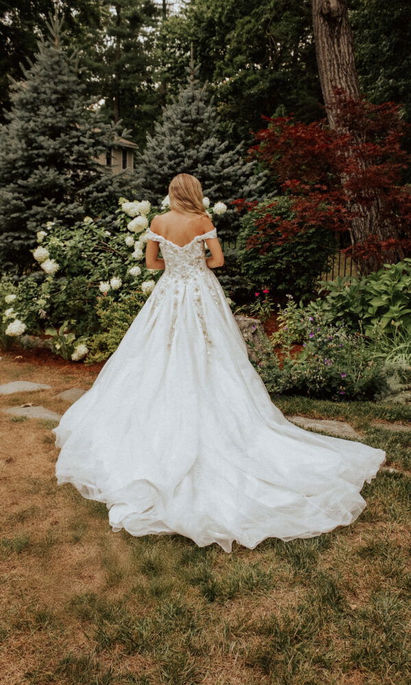 Die Braut in einem eleganten Brautkleid posiert von hinten in einem grünen Garten, die Schleppe ihres Kleides breitet sich auf dem Boden aus.