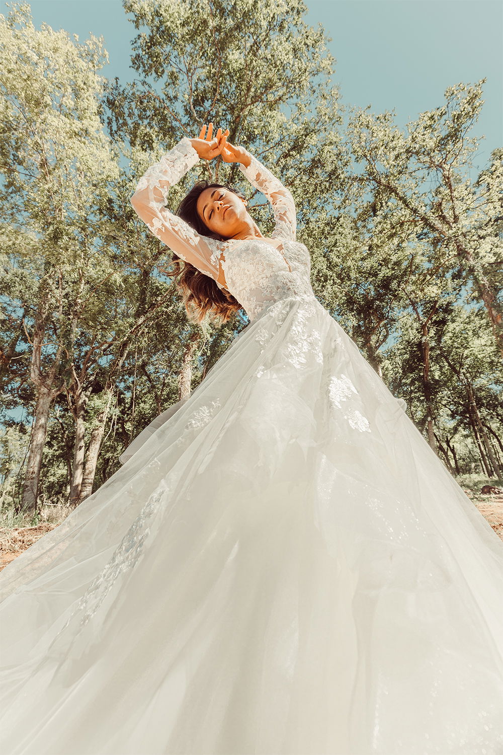 In einem waldigen Umfeld posiert die Braut in einem Kleid mit floralen Spitzenmustern, die Arme erhoben, umgeben von Natur.