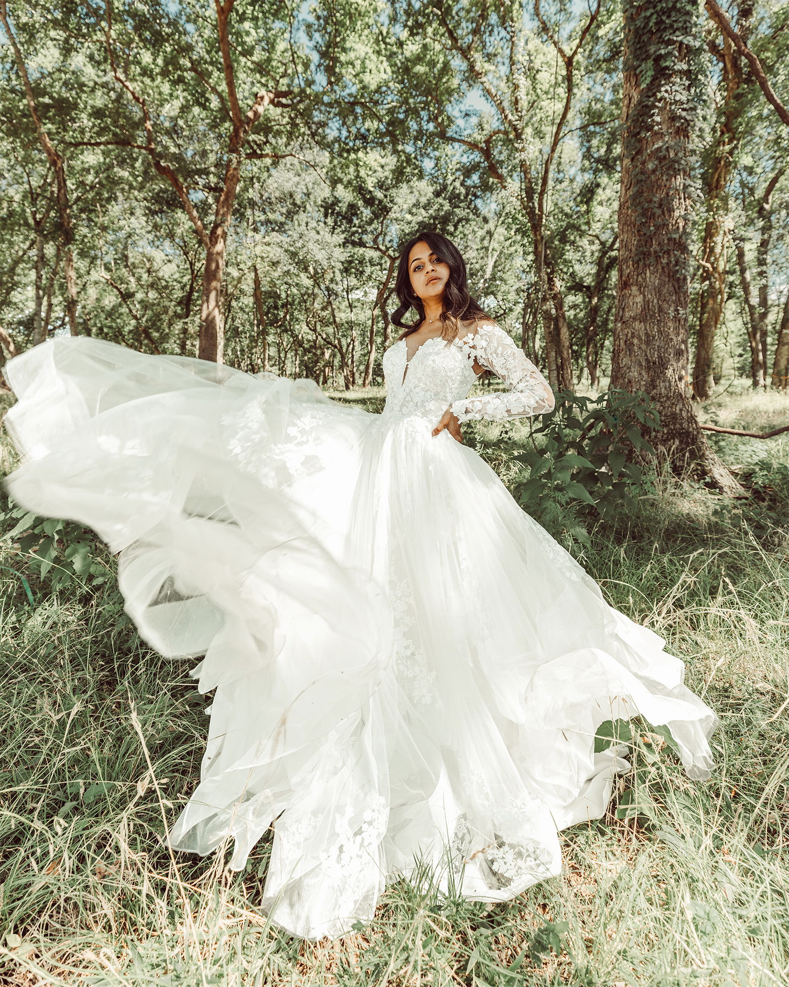 Inmitten von Bäumen und Gras steht die Braut in einem Kleid mit floraler Spitze, die Schleppe des Kleides weht dramatisch zur Seite.