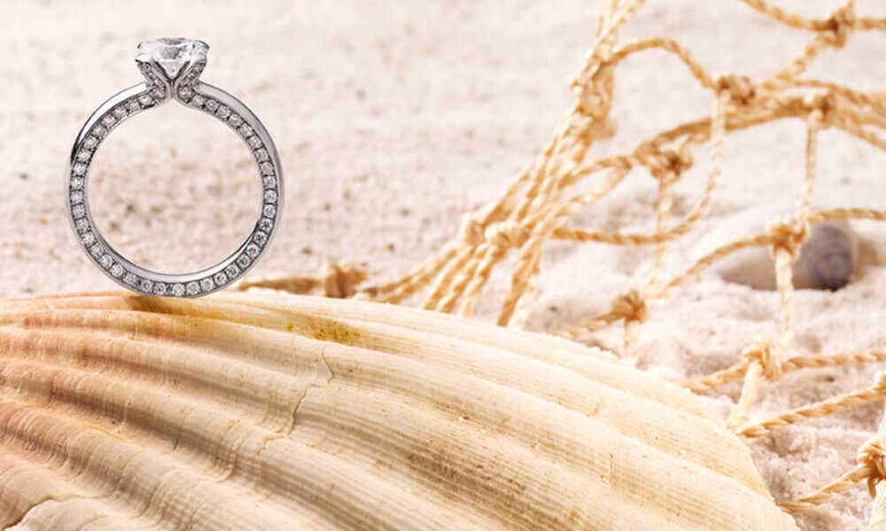 Ein funkelnder Verlobungsring mit Diamanten, der elegant auf einer Muschel am Strand platziert ist. Im Hintergrund sind ein Netz und der sandige Boden zu sehen, die eine romantische, maritime Atmosphäre schaffen.
