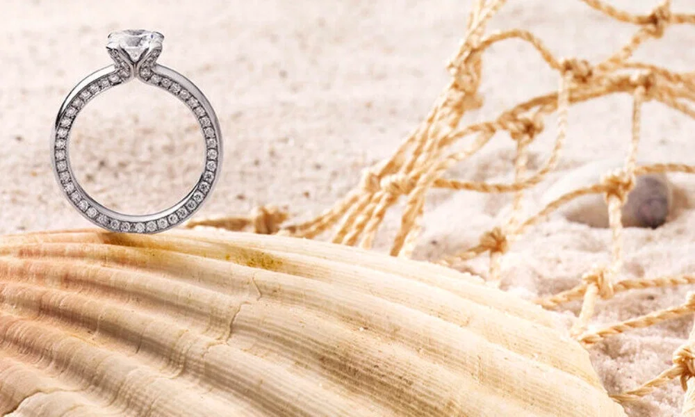 Ein funkelnder Verlobungsring mit Diamanten, der elegant auf einer Muschel am Strand platziert ist. Im Hintergrund sind ein Netz und der sandige Boden zu sehen, die eine romantische, maritime Atmosphäre schaffen.