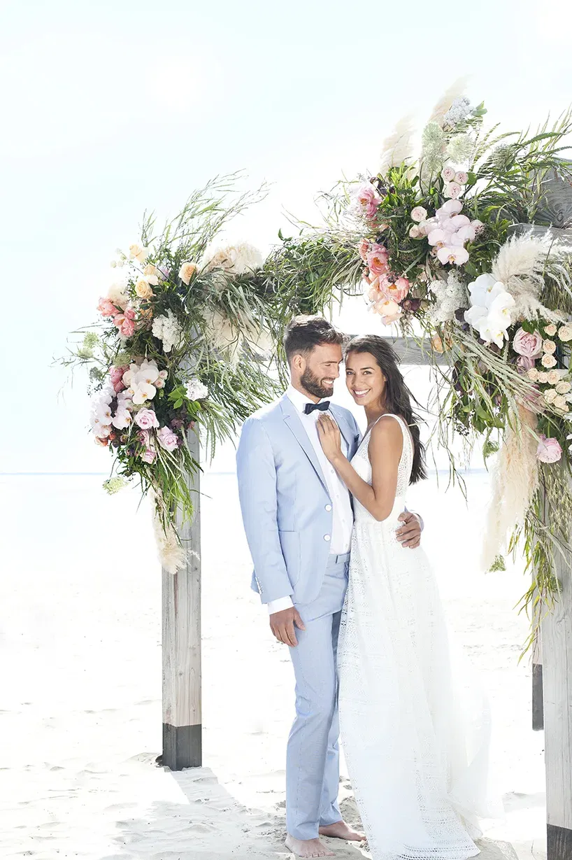 Ein Brautpaar posiert vor einem mit Blumen geschmückten Bogen am Strand, die Braut im romantischen Kleid, der Bräutigam im hellblauen Anzug.