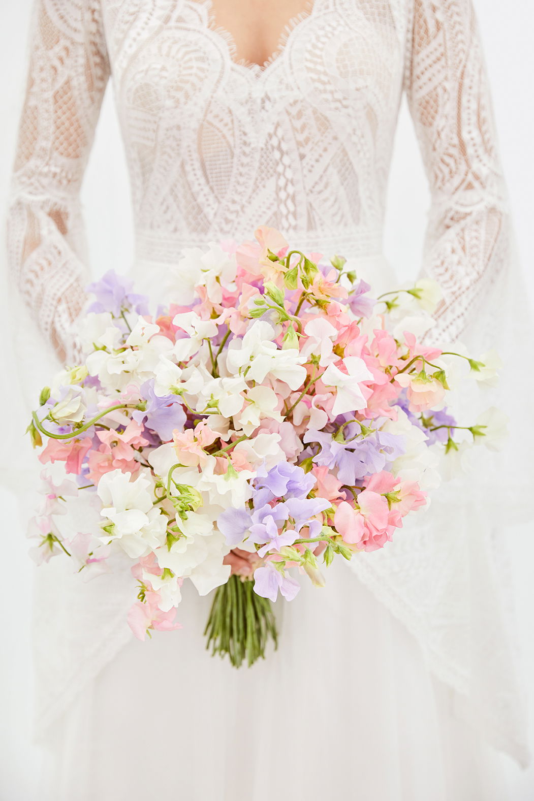 Ein pastellfarbener Brautstrauß mit Blüten in Rosa, Lila und Weiß, gehalten von einer Braut im feinen Spitzenhochzeitskleid.