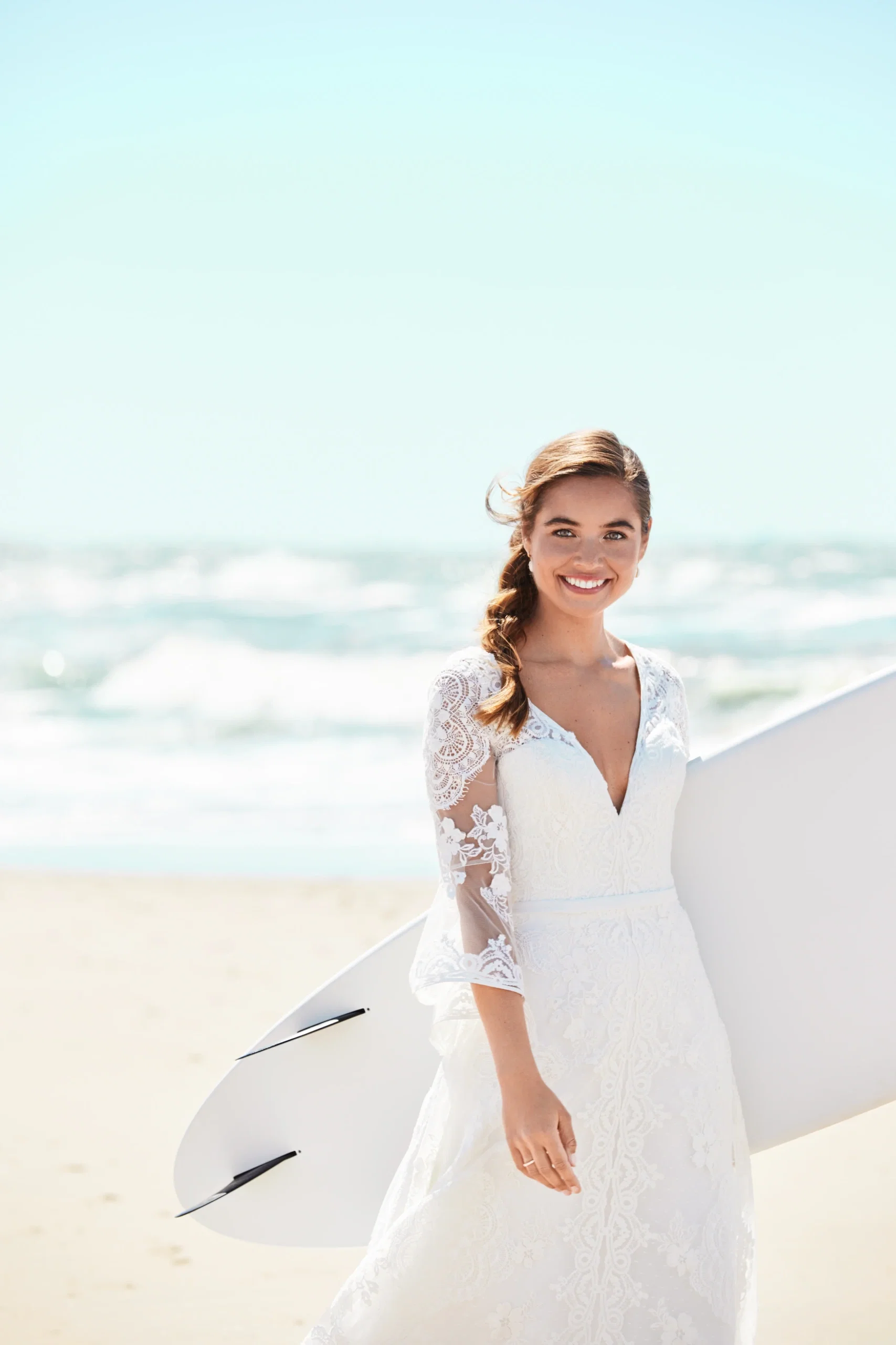 Junge Braut im Boho-Stil mit Surfbrett am Strand, in einem Spitzenkleid und mit entspanntem Lächeln vor der Meereskulisse.