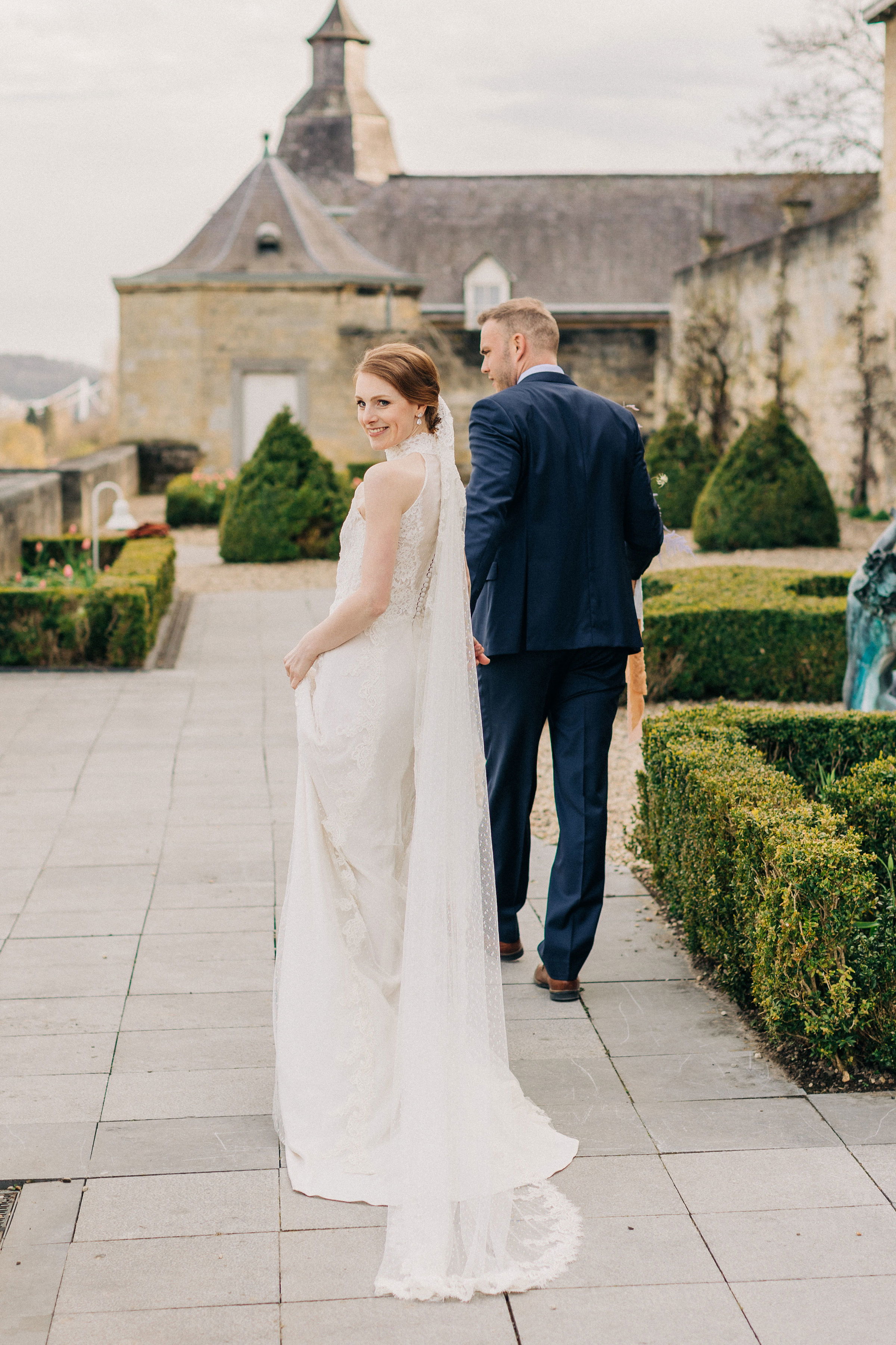 Ein Brautpaar geht Hand in Hand durch eine gepflegte Gartenanlage vor einem historischen Gebäude. Die Braut trägt ein elegantes, bodenlanges Hochzeitskleid mit langer Spitze, während der Bräutigam einen dunkelblauen Anzug trägt. Die Braut blickt lächelnd über die Schulter zurück.