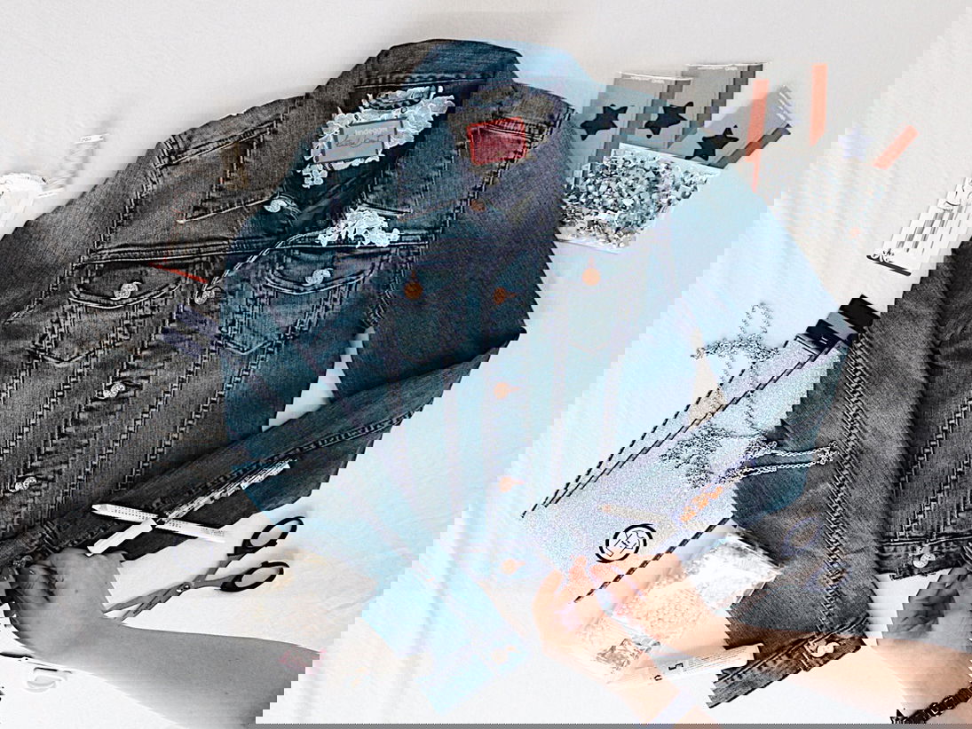 Eine flach gelegte Jeansjacke wird von verschiedenen DIY-Utensilien wie Glitzerfolie, Perlen, Nadel und Faden umgeben. Hände arbeiten an der Jacke und verleihen ihr eine persönliche Note.