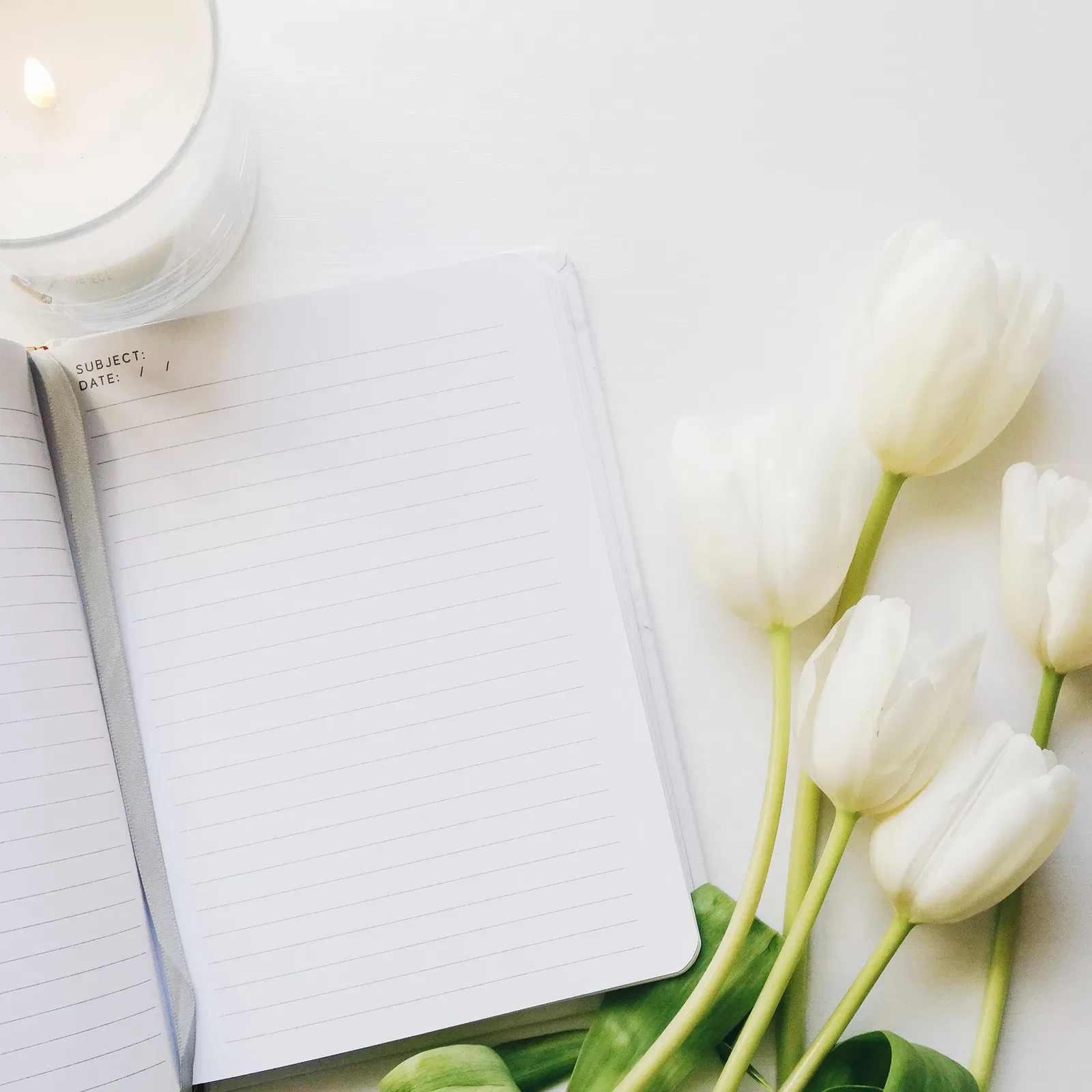 Ein geöffnetes Notizbuch mit linierten Seiten auf einem weißen Untergrund, daneben ein angezündetes Teelicht in einem Glasbehälter. Rechts vom Notizbuch liegen mehrere weiße Tulpen, deren Köpfe zum Notizbuch hin geneigt sind, was einen Eindruck von Frische und Einfachheit vermittelt und zum Schreiben oder Planen in einer ruhigen, inspirierenden Umgebung einlädt.