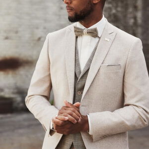 Ein stilvoller Bräutigam in einem hellbeigen Boho-Hochzeitsanzug mit Weste und Fliege, der selbstbewusst im Freien steht und eine Sonnenbrille trägt.