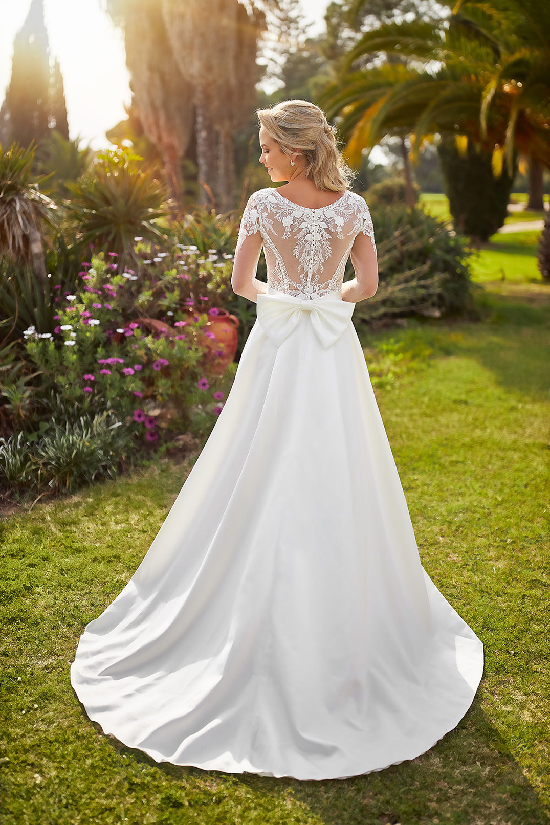 Braut in einem Kleid mit feinem Spitzenmuster und großer Schleife am Rücken. Sie posiert in einem Garten voller Blumen und Palmen.