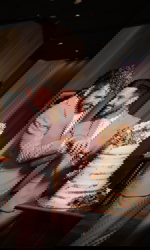 Gleichgeschlechtliches Paar in abgestimmten Anzügen beim Anschneiden der Hochzeitstorte, beide strahlen vor Glück.