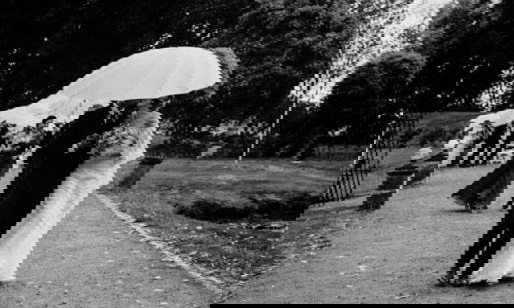 Das Brautpaar geht unter einem weißen Schirm entlang eines Waldwegs, liebevoll Arm in Arm, während es leicht regnet und Gäste im Hintergrund folgen.