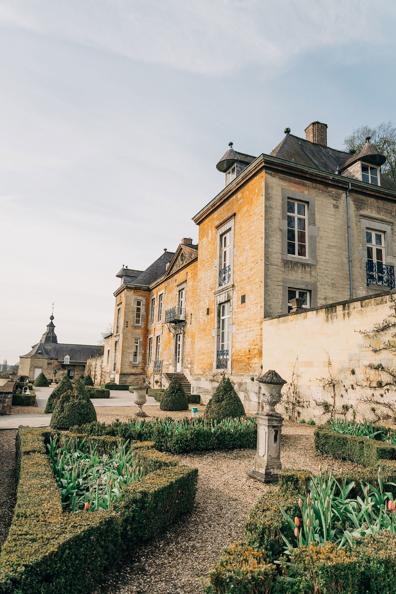 Ein prachtvolles Herrenhaus aus hellem Stein mit barockem Garten, dekoriert mit Buchsbaumhecken und Skulpturen, im Licht der Abendsonne.