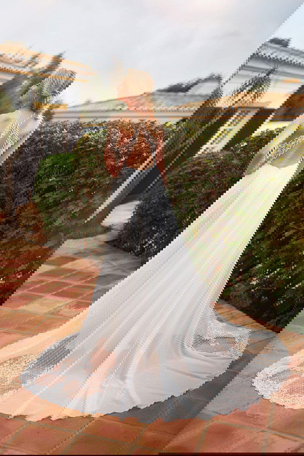 Braut in einem eleganten Kleid mit offenem Rückendetail und Spitzenakzenten. Sie steht auf einer Terrasse mit grüner Hecke und mediterranem Flair.