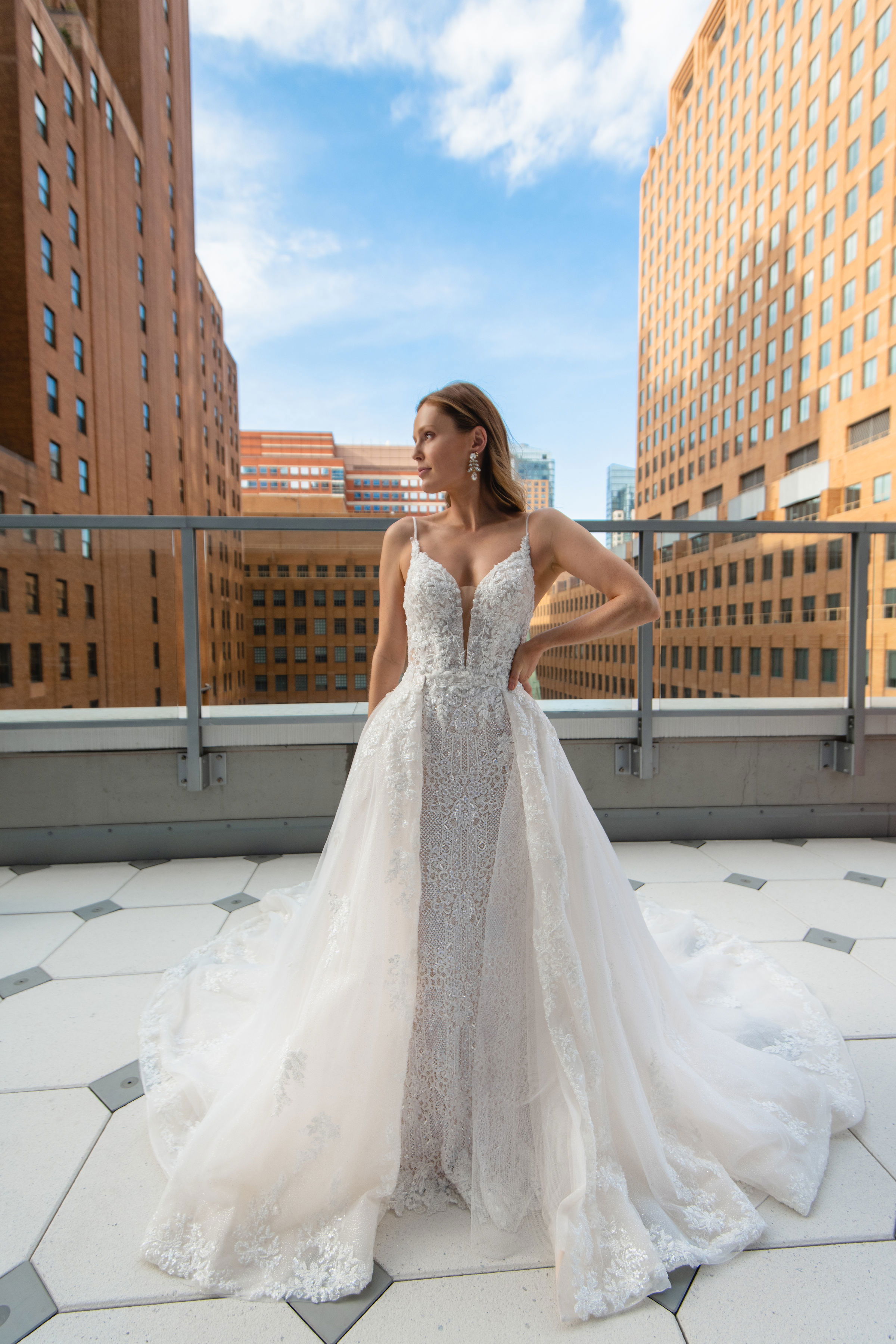 Auf einer urbanen Dachterrasse steht die Braut in einem Spitzenkleid, das detaillierte Muster und eine lange Schleppe aufweist. Im Hintergrund sind hohe Gebäude sichtbar, was dem Bild eine moderne Atmosphäre verleiht.