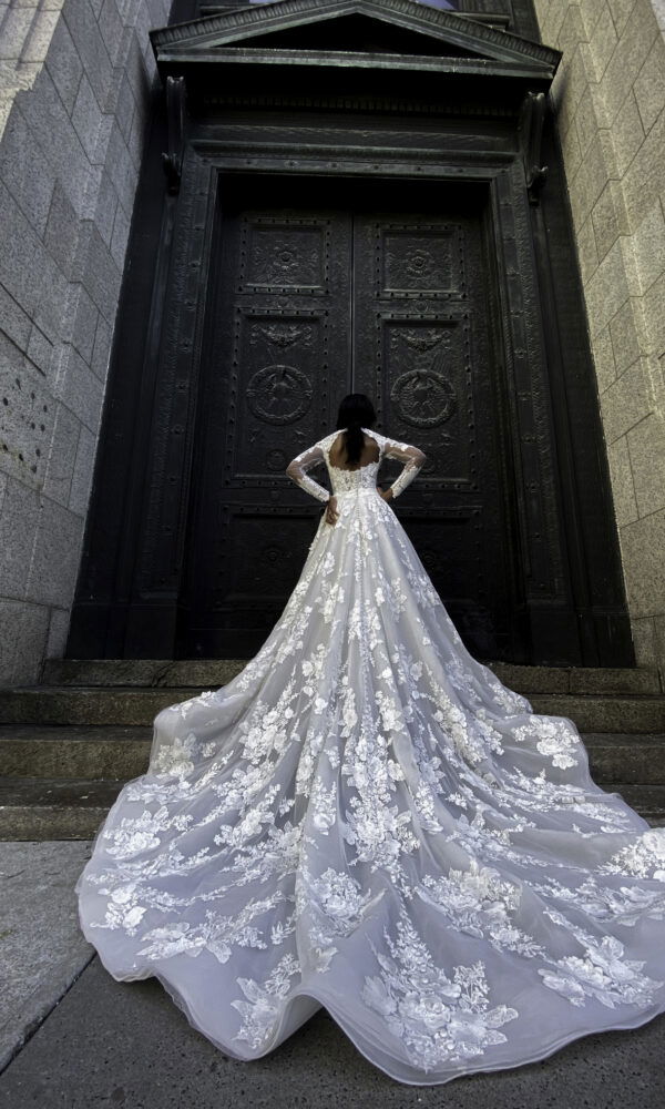 Elegantes Brautkleid mit detaillierter Spitze und einer majestätischen Schleppe, getragen von einer Braut, die vor einer imposanten Tür steht und einen kraftvollen Look präsentiert.
