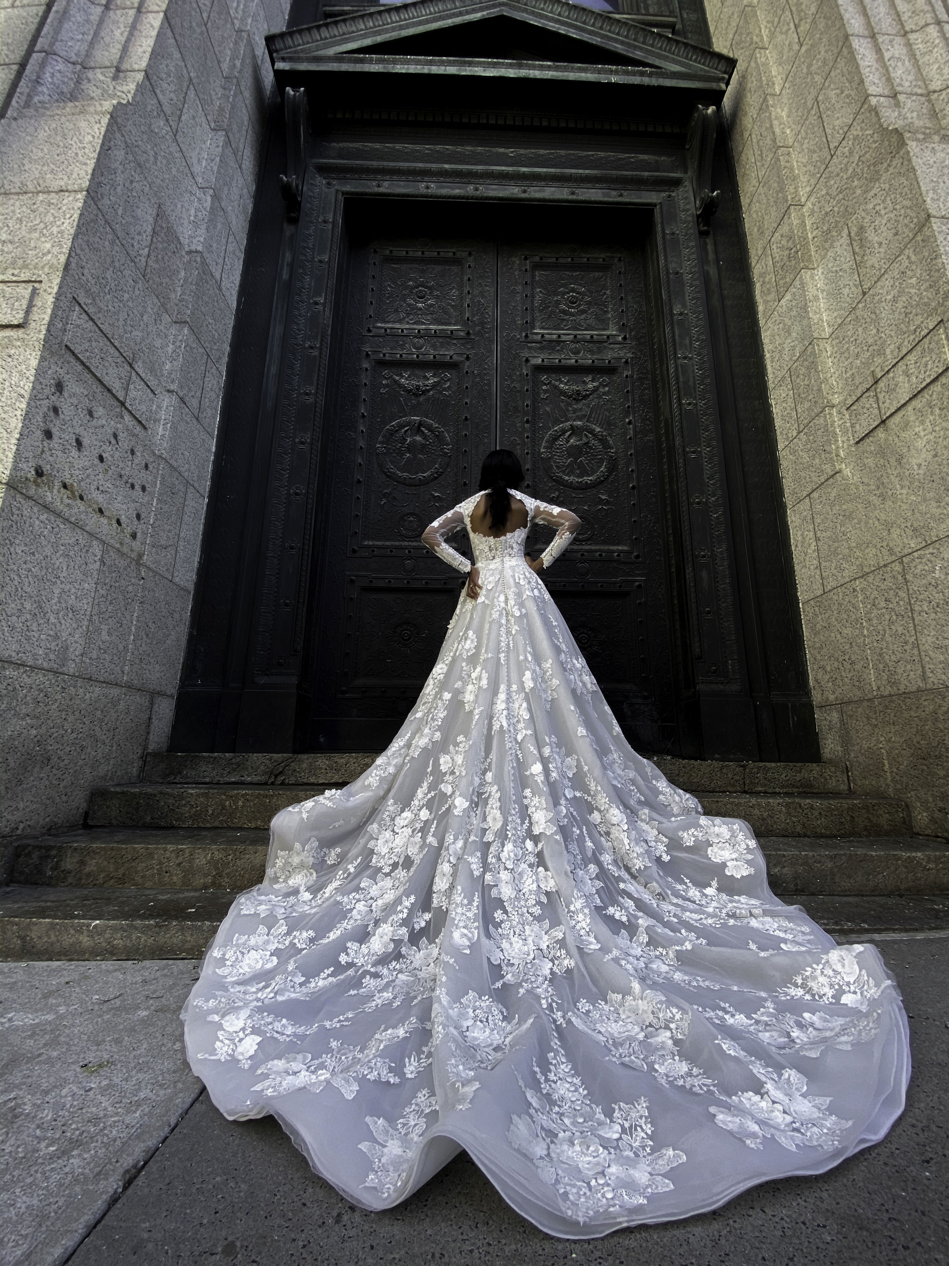 Elegantes Brautkleid mit detaillierter Spitze und einer majestätischen Schleppe, getragen von einer Braut, die vor einer imposanten Tür steht und einen kraftvollen Look präsentiert.