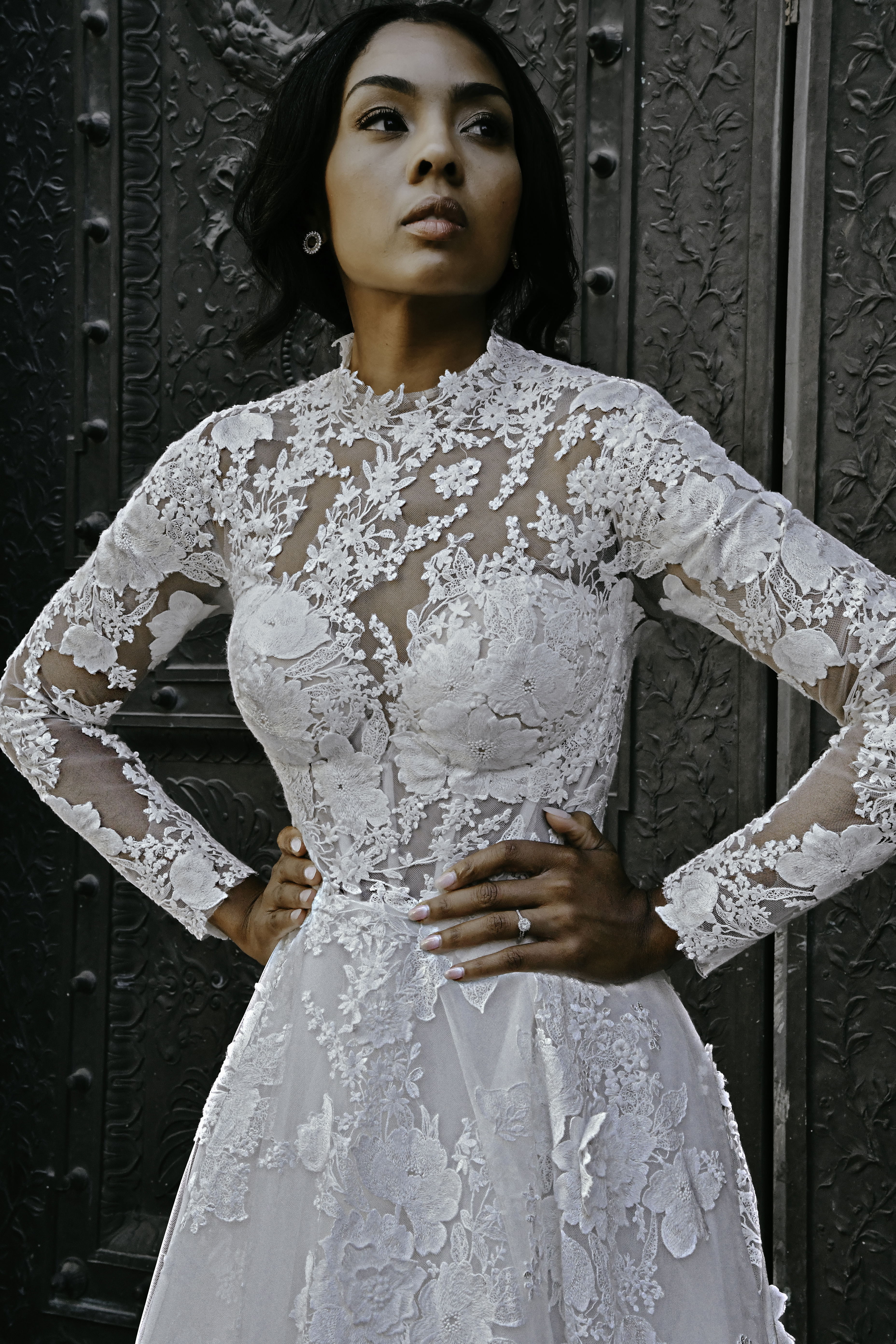 Braut mit langärmligem Spitzenkleid und floralem Muster im Oberteil, die vor einer dekorativen dunklen Tür posiert und stolz in die Ferne schaut.