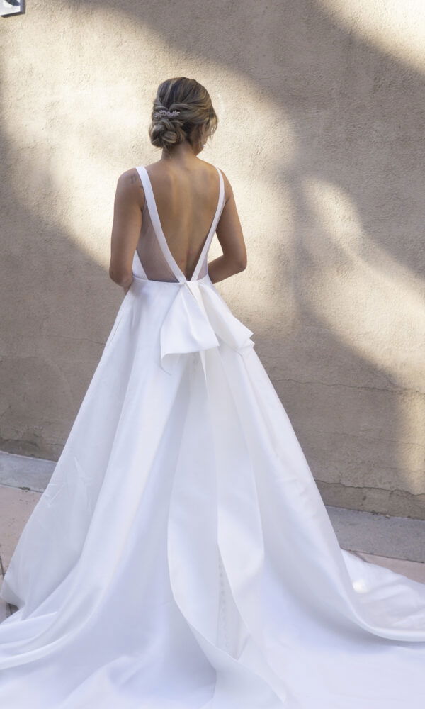 Eine Frau im eleganten weißen Brautkleid mit offenem Rücken und Schleifendetail, die vor einer neutralen Wand mit Schatteneffekten steht.