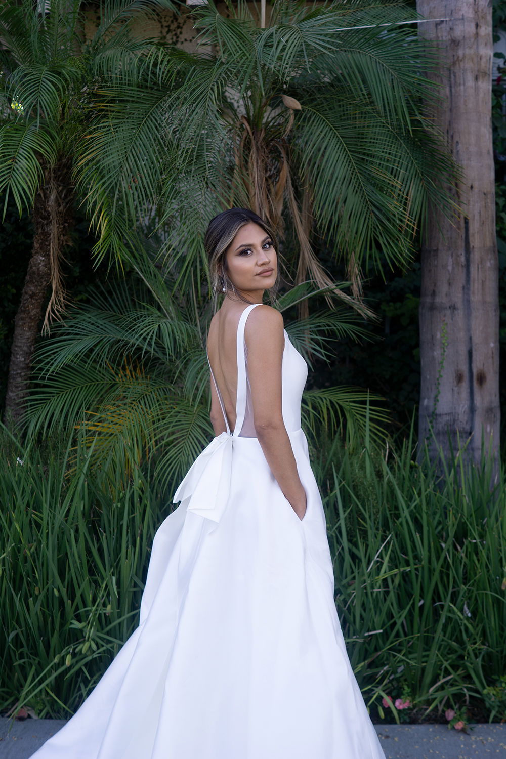 Eine Frau im schlichten weißen Brautkleid mit offenem Rücken und Schleifenakzent, stehend vor einer Wand aus Palmenblättern.