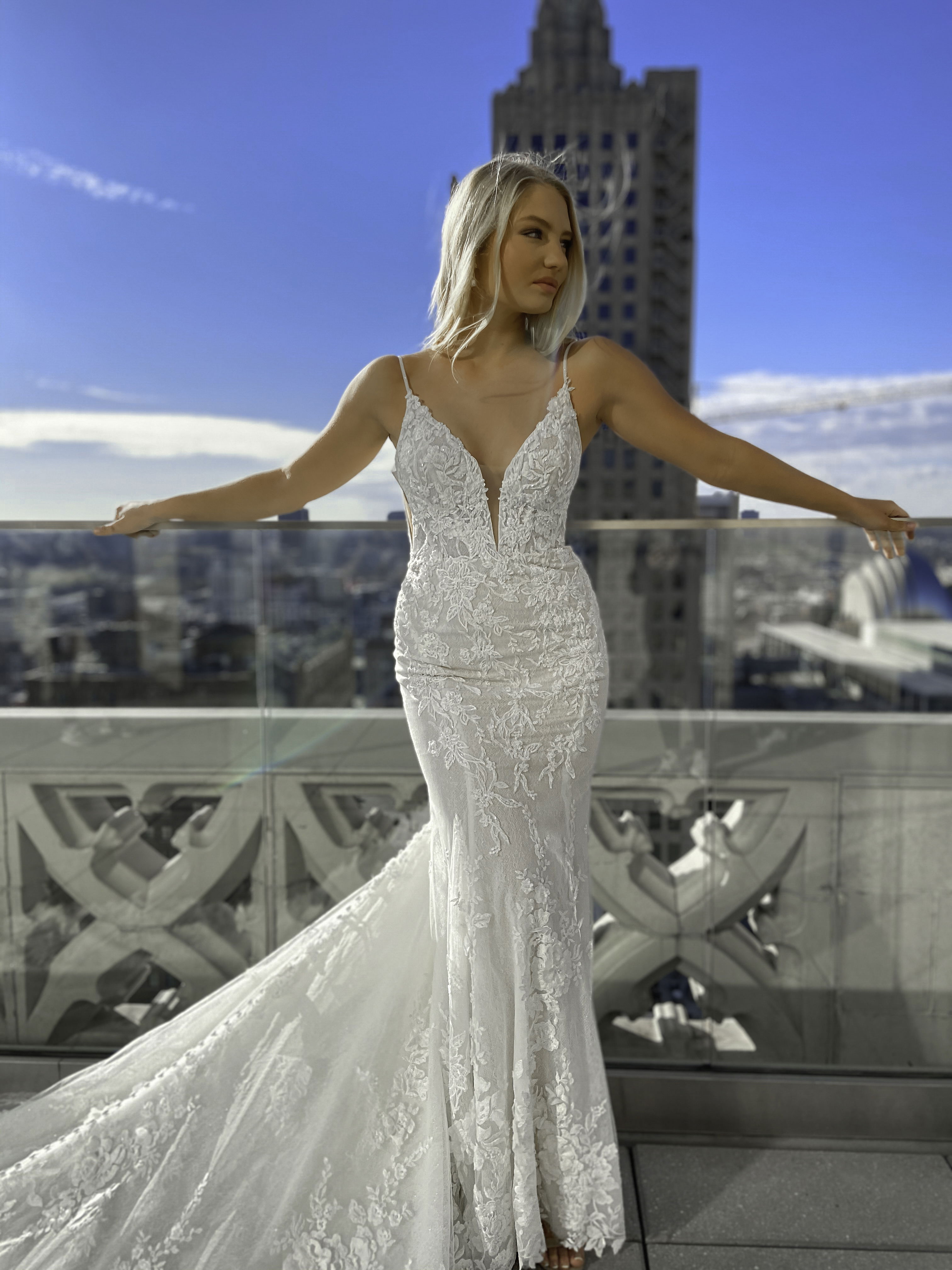 In einem spitzenverzierten Hochzeitskleid mit tiefem Ausschnitt breitet die Braut ihre Arme auf einem Balkon aus und genießt den Ausblick über die Stadt.