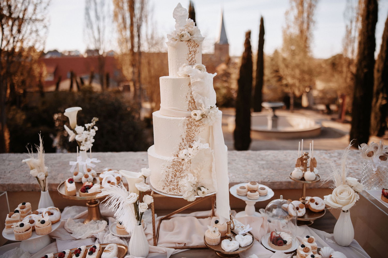 Weißer Desserttisch im Freien mit einer kunstvollen Hochzeitstorte, kleinen Cupcakes und Blumen als perfekte Ergänzung für eine stilvolle Hochzeit.