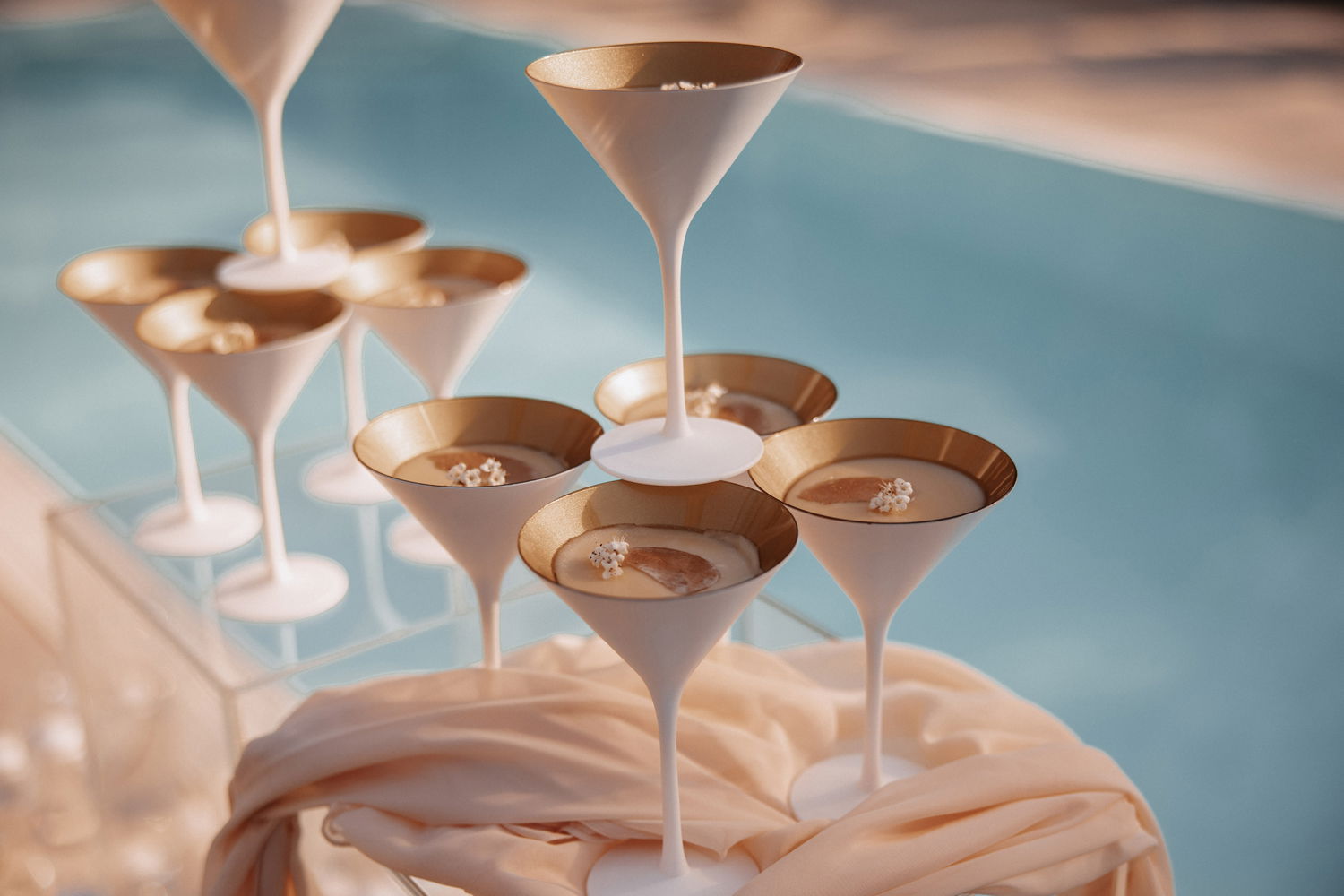 Elegant platzierte Martini-Gläser mit mattem Finish und Goldrand spiegeln sich sanft am Pool, arrangiert auf einem weichen, pastellfarbenen Tuch – ein Highlight für Hochzeitsdekorationen.