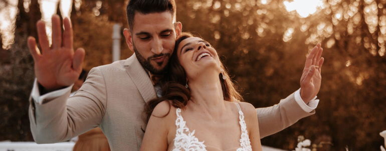 Lächelndes Brautpaar bei Sonnenuntergang, sie trägt ein Spitzenkleid, er einen hellen Anzug, beide genießen ihren besonderen Tag.