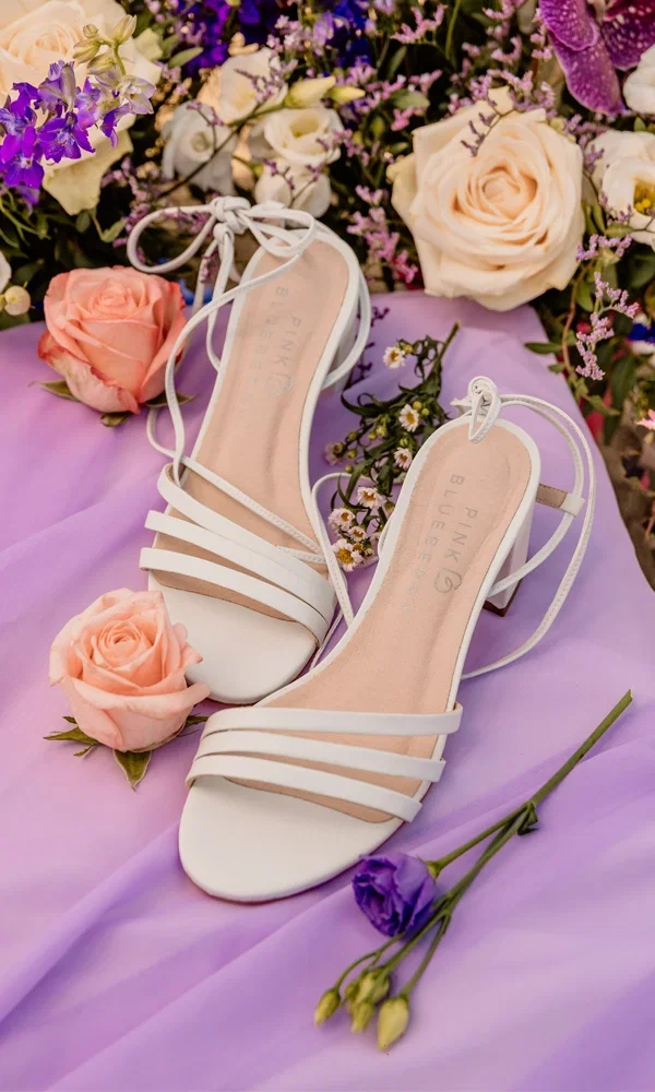 Zwei weiße, minimalistische Hochzeitssandalen mit eleganten Riemchen liegen auf einem lila Stoff, umgeben von einem Blumenarrangement aus Rosen und anderen farbenfrohen Blüten. Die Sandalen haben einen modernen, schlichten Stil, ideal für eine sommerliche Hochzeit im Freien. Das Bild wird von sanften floralen Akzenten umrahmt und erzeugt eine harmonische, romantische Atmosphäre.