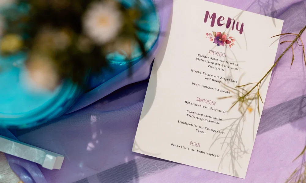 Ein handgeschrieben wirkendes Menü auf hellem Papier, verziert mit einem blumigen Design am oberen Rand, liegt auf einem violettem Tuch. Neben der Karte sind verschwommene Elemente wie ein blauer Teller und Blumen zu erkennen. Die Speisekarte umfasst Salate, Hauptgerichte wie Schollenfilet mit Champagnersauce und ein Erdbeer-Panna-Cotta-Dessert.