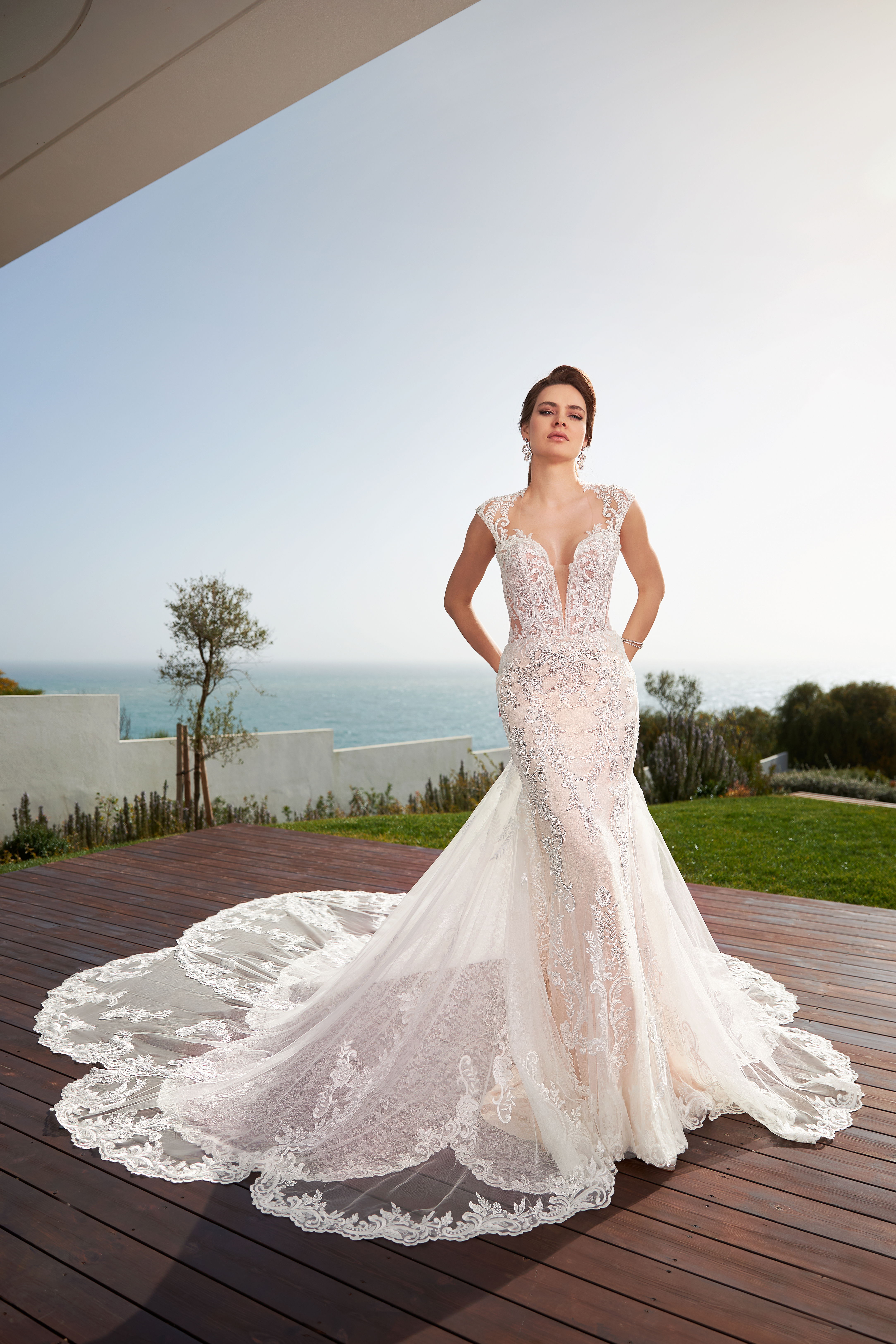 Brautkleid mit detaillierter Spitze und langer Schleppe auf einer Terrasse am Meer. Die Braut posiert selbstbewusst und genießt den Ausblick.