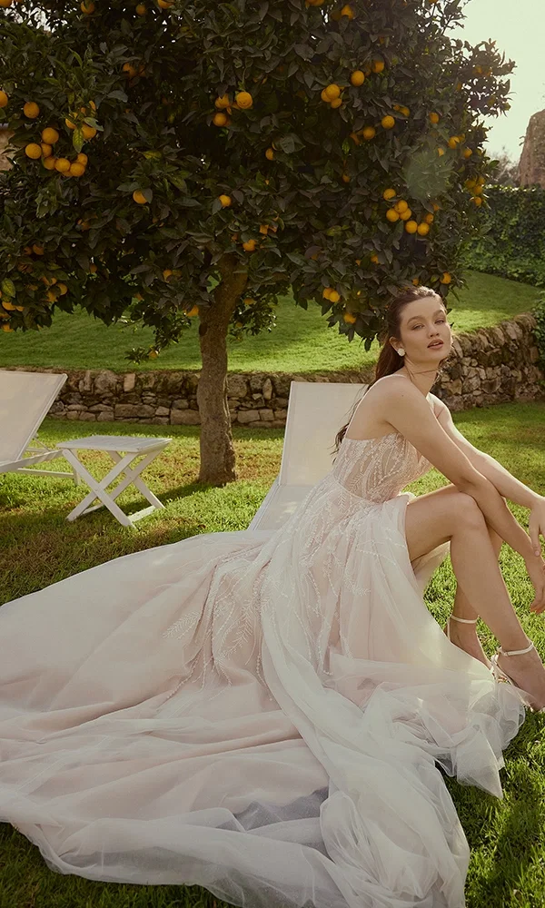 Braut in einem modernen, rosafarbenen Hochzeitskleid, sitzt entspannt auf einer Wiese unter einem Orangenbaum, umgeben von einem sommerlichen Gartenambiente.