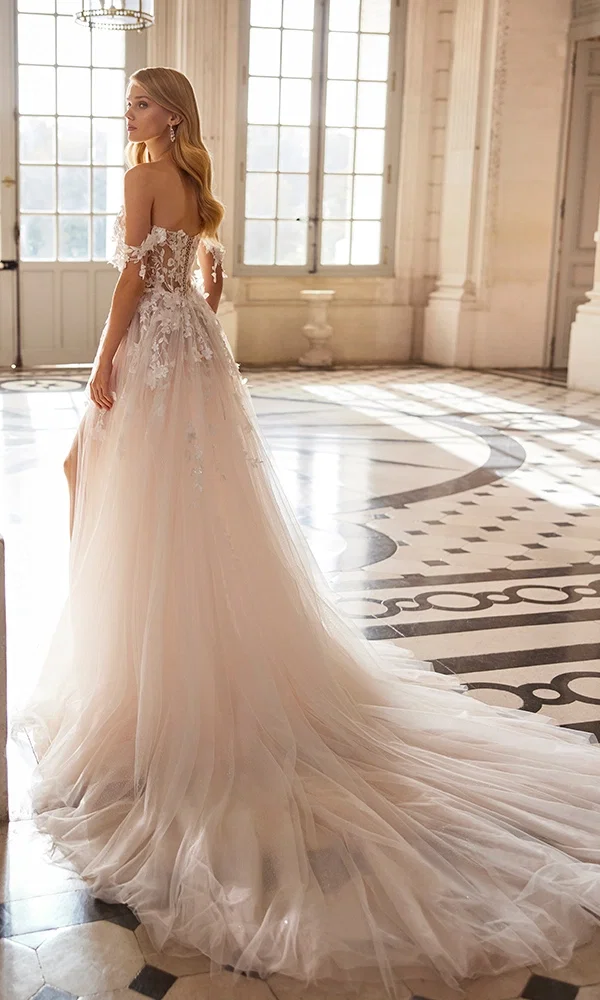 Braut in einem romantischen, rosafarbenen Hochzeitskleid mit zarten Spitzendetails, posiert in einem lichtdurchfluteten Raum mit Marmorboden und großen Fenstern, die auf einen malerischen Garten blicken.