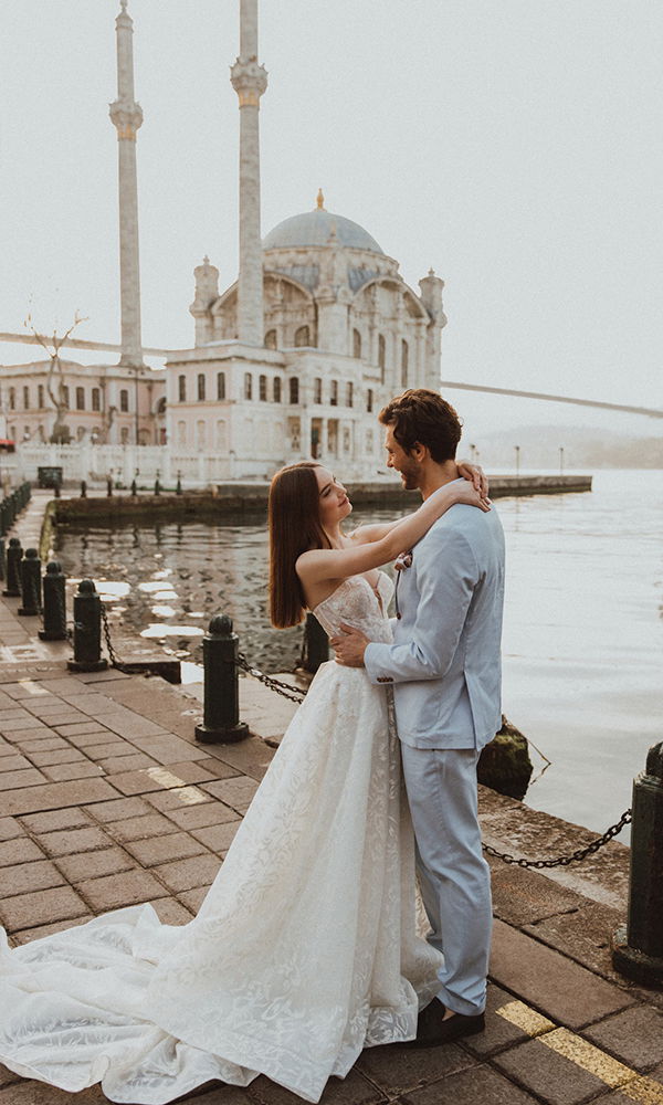 Verliebtes Paar in Hochzeitskleidung an der Uferpromenade vor einer Moschee und Brücke. Die Braut trägt ein elegantes Spitzenkleid mit Schleppe, der Bräutigam einen hellen Anzug – ein Moment voller Zärtlichkeit.