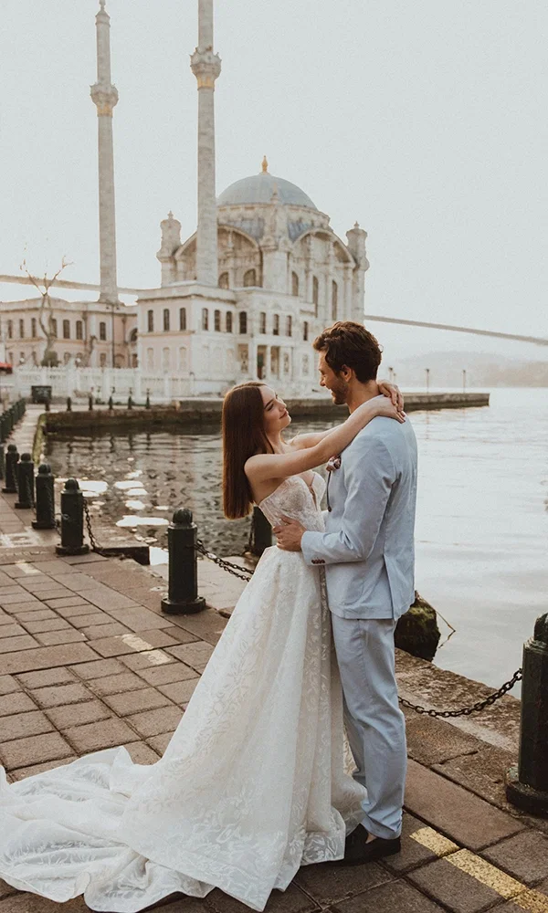 Verliebtes Paar in Hochzeitskleidung an der Uferpromenade vor einer Moschee und Brücke. Die Braut trägt ein elegantes Spitzenkleid mit Schleppe, der Bräutigam einen hellen Anzug – ein Moment voller Zärtlichkeit.