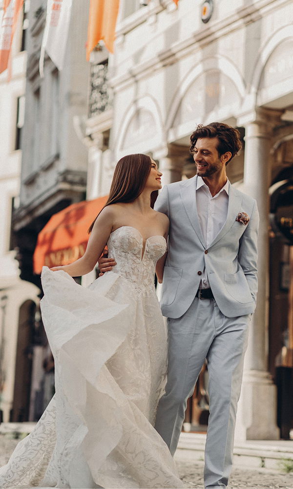 Verliebtes Paar in Hochzeitskleidung schlendert durch die Stadt. Die Braut im trägerlosen Brautkleid mit verspielten Details, der Bräutigam im hellen Anzug – ein Moment der Verbundenheit und Freude in einer städtischen Kulisse.