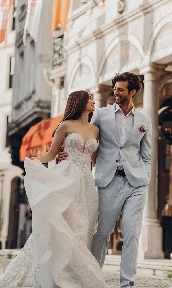Verliebtes Paar in Hochzeitskleidung schlendert durch die Stadt. Die Braut im trägerlosen Brautkleid mit verspielten Details, der Bräutigam im hellen Anzug – ein Moment der Verbundenheit und Freude in einer städtischen Kulisse.