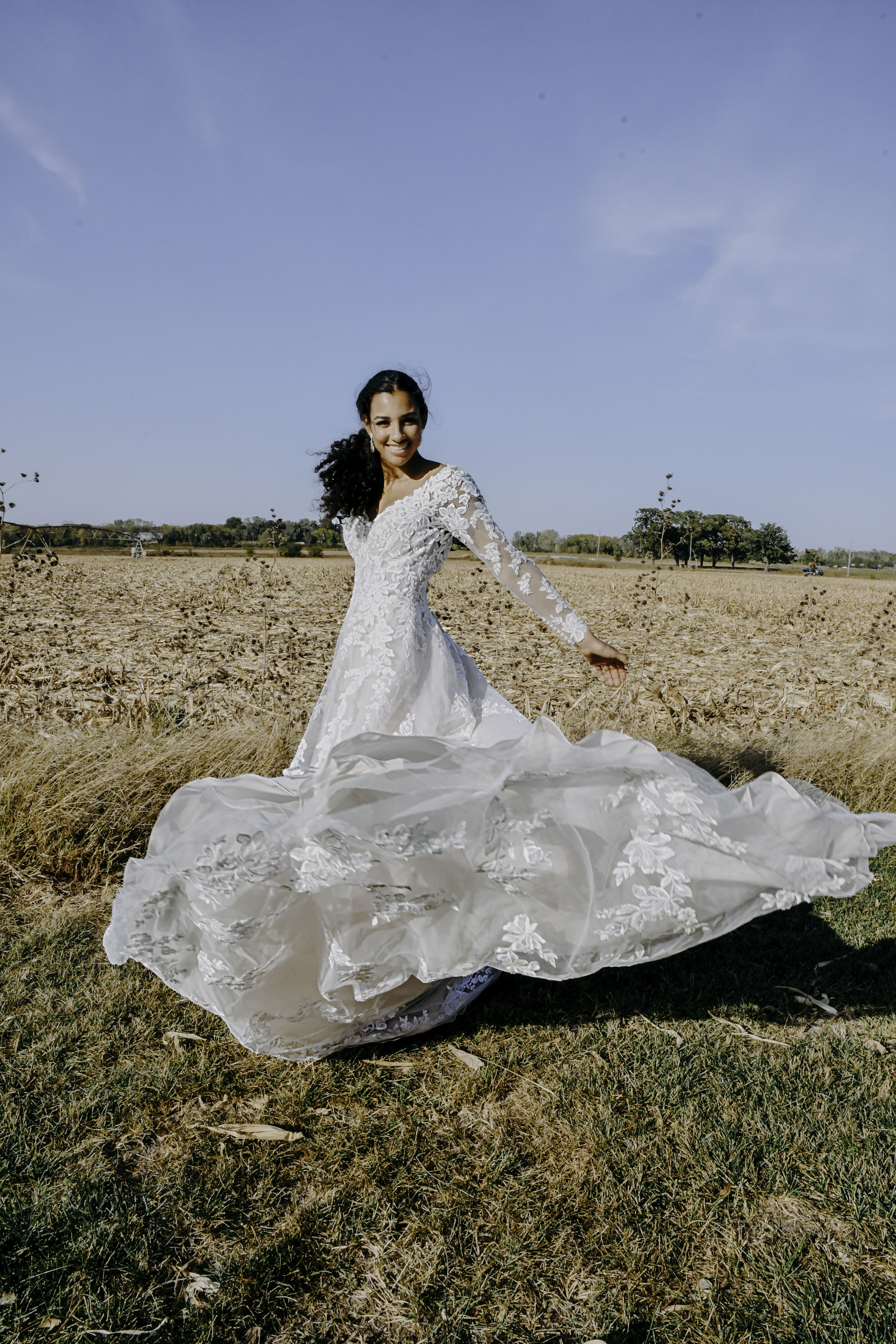 Auf einem weiten Feld dreht sich eine Braut in einem detailreichen Kleid mit Spitze, lächelnd in die Kamera, mit wehender Schleppe.