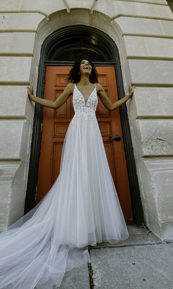 Elegante Braut im langen, weißen Hochzeitskleid mit Spitzendetails und Schleppe, posiert lächelnd vor einer roten Tür.