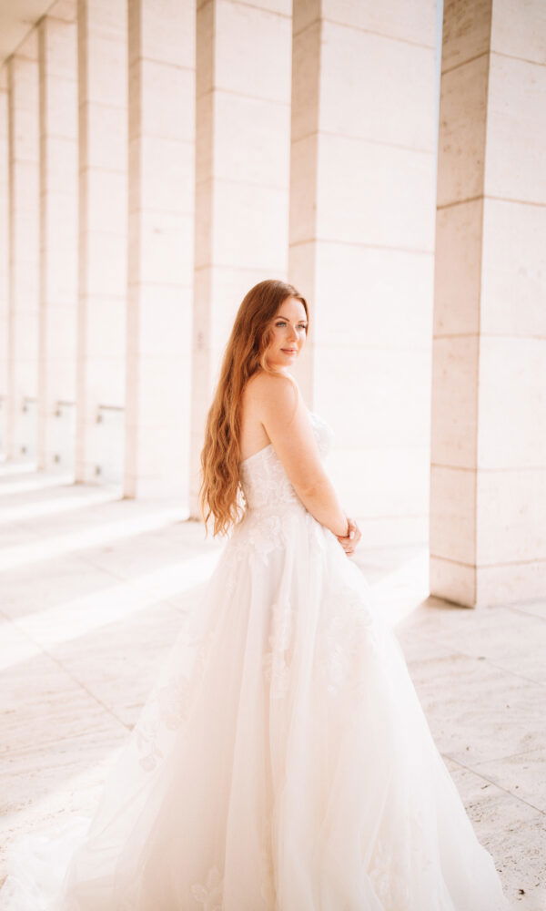Braut im ärmellosen Kleid in einem modernen, hellen Raum mit Marmorsäulen. Ihr langes, rotes Haar fällt weich über ihren Rücken.