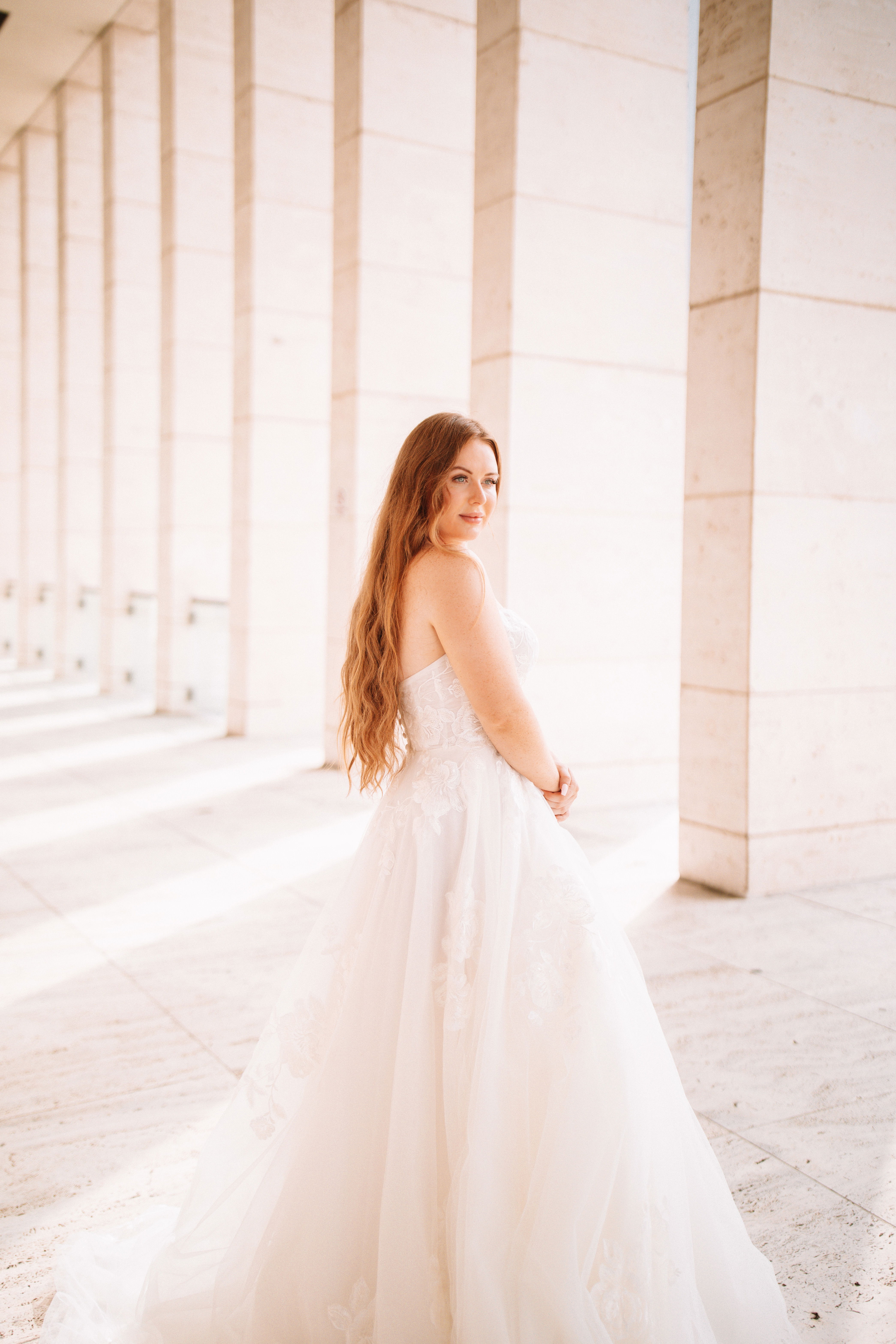 Braut im ärmellosen Kleid in einem modernen, hellen Raum mit Marmorsäulen. Ihr langes, rotes Haar fällt weich über ihren Rücken.