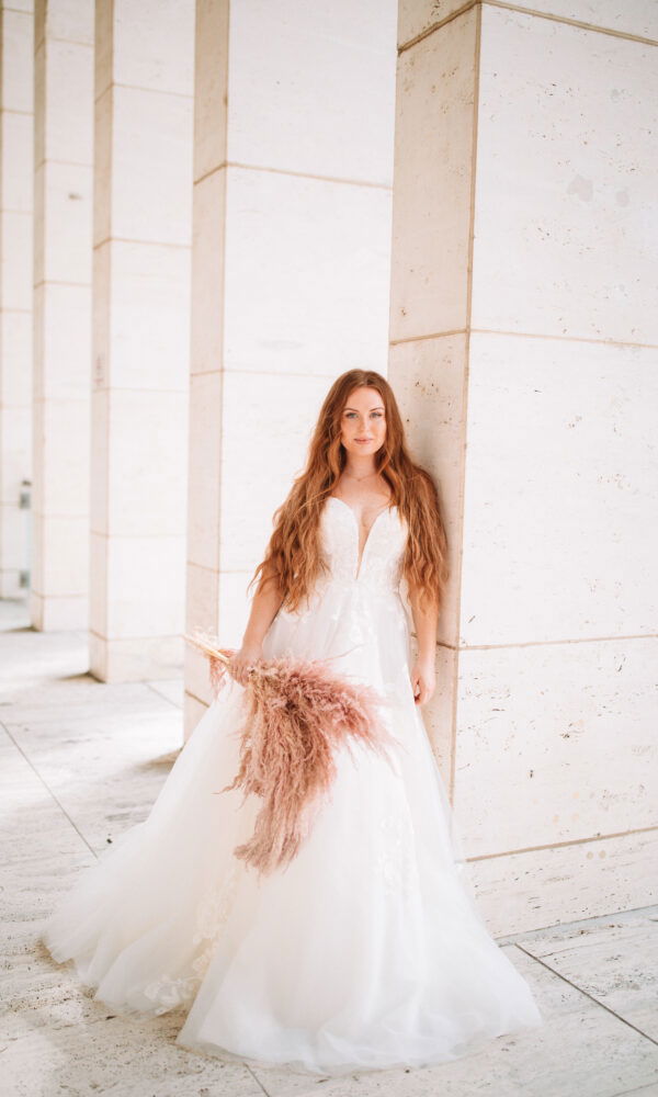 Braut in einem weißen Kleid lehnt an einer Steinwand und blickt direkt in die Kamera. Ihre langen, rotbraunen Haare fallen über die Schultern, und sie hält einen Strauß aus beige-rosa Trockenblumen. Die minimalistische Umgebung schafft einen modernen Look.