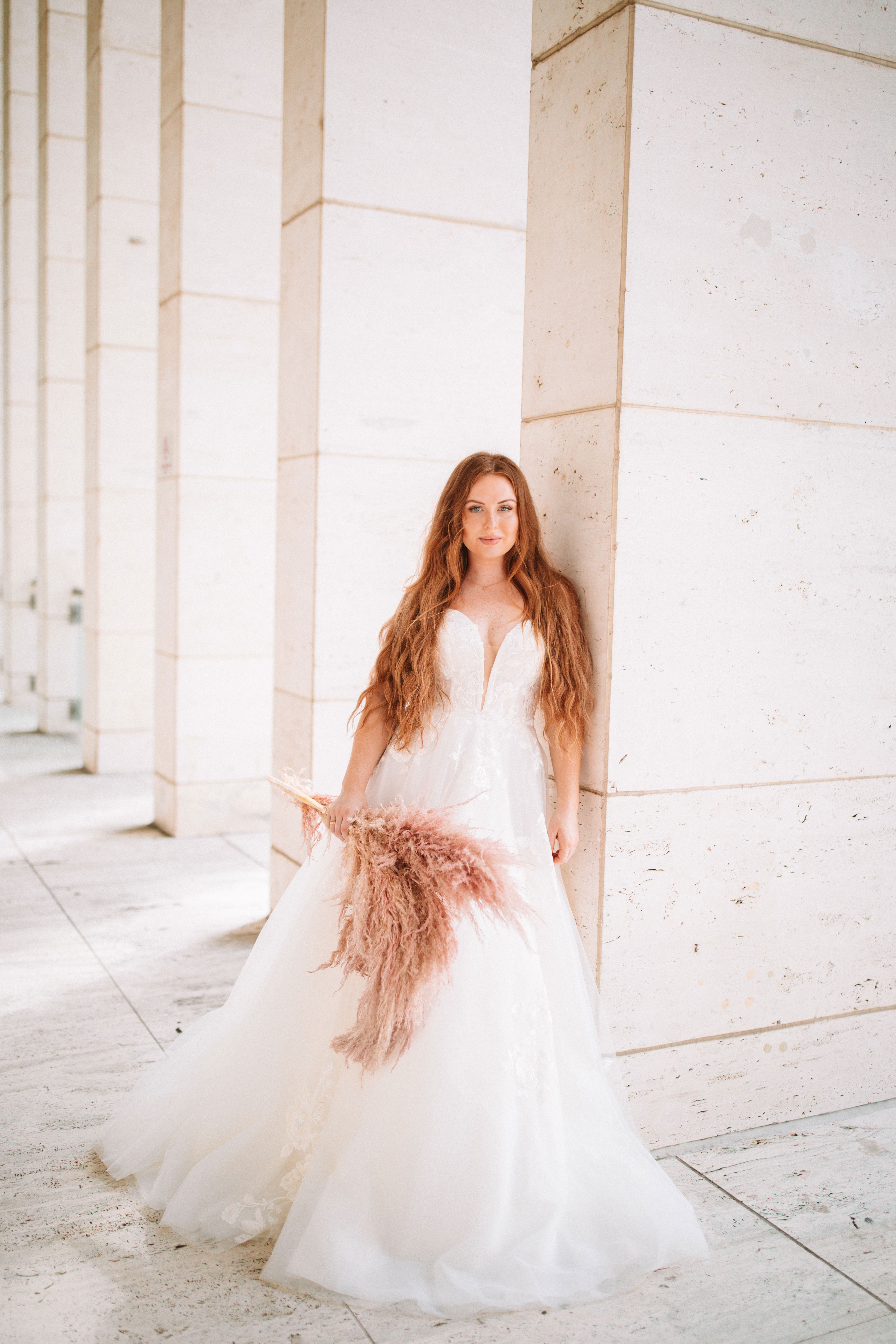 Braut in einem weißen Kleid lehnt an einer Steinwand und blickt direkt in die Kamera. Ihre langen, rotbraunen Haare fallen über die Schultern, und sie hält einen Strauß aus beige-rosa Trockenblumen. Die minimalistische Umgebung schafft einen modernen Look.