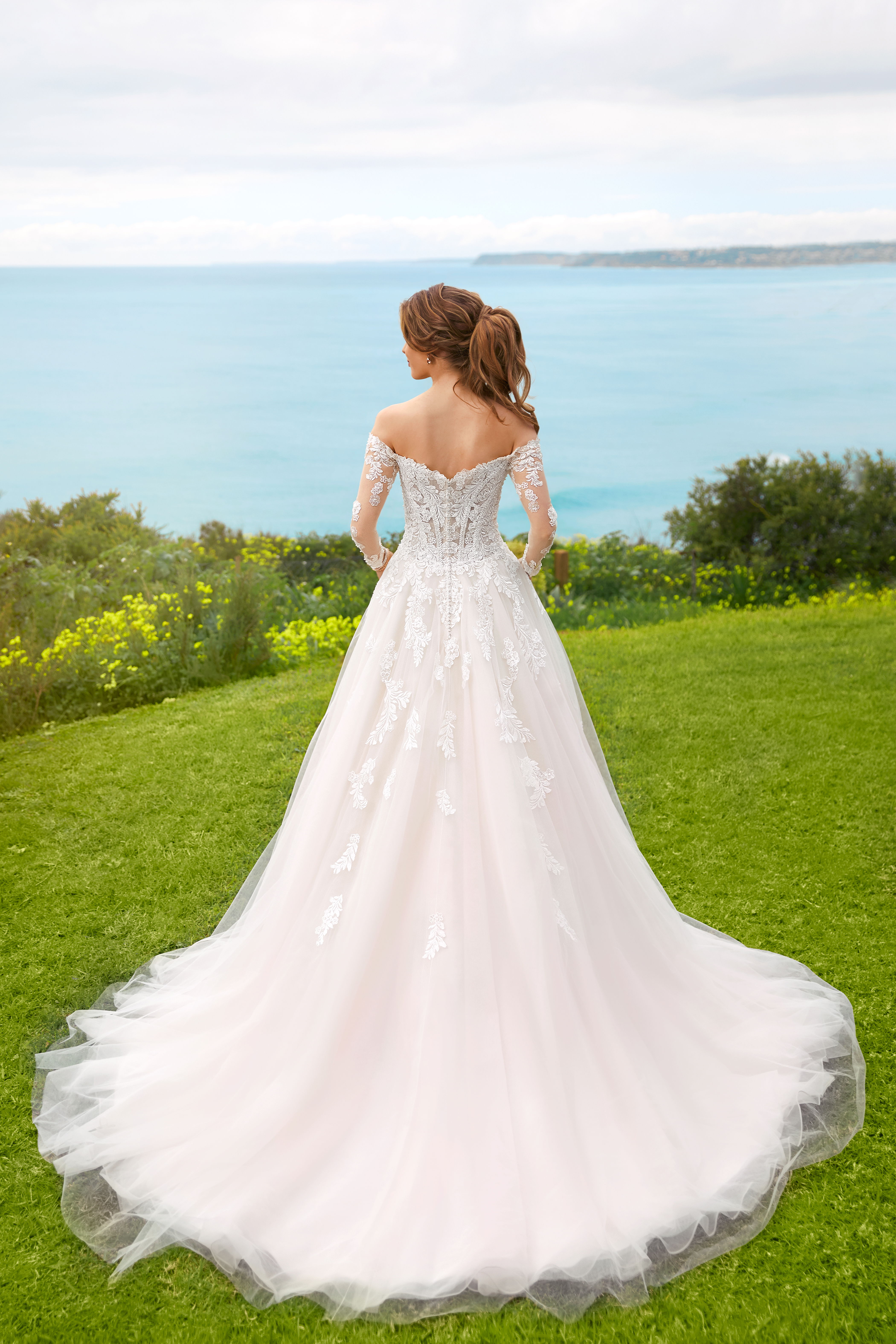 Braut in einem trägerlosen Ballkleid aus Tüll und Spitze auf einer Wiese, die den Blick auf das Meer und die Landschaft im Hintergrund genießt.
