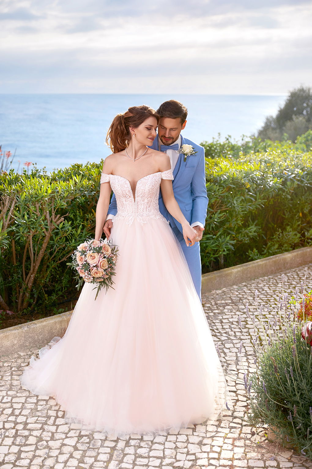 Braut und Bräutigam genießen einen romantischen Moment am Meer. Die Braut trägt ein zartes Kleid mit Rosenstrauß, der Bräutigam einen blauen Anzug.