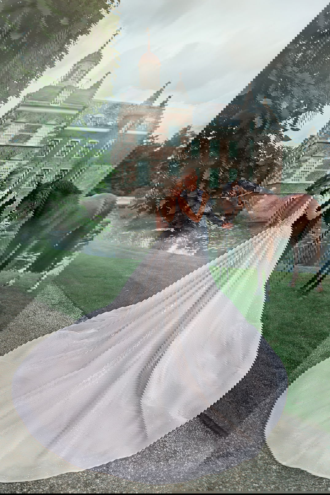 Braut und Bräutigam posieren vor einem Herrenhaus mit einem Pferd. Die Braut trägt ein langes Kleid mit Schleppe, der Bräutigam hält das Pferd.